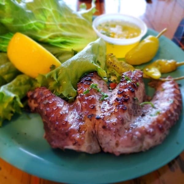 Grilled octopus with fresh lettuce, a lemon wedge, and pickled peppers on a teal plate.